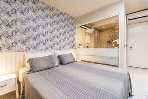 Quarto moderno com uma cama grande, uma mesa de cabeceira, banheiro com chuveiro e um espelho refletindo o banheiro.