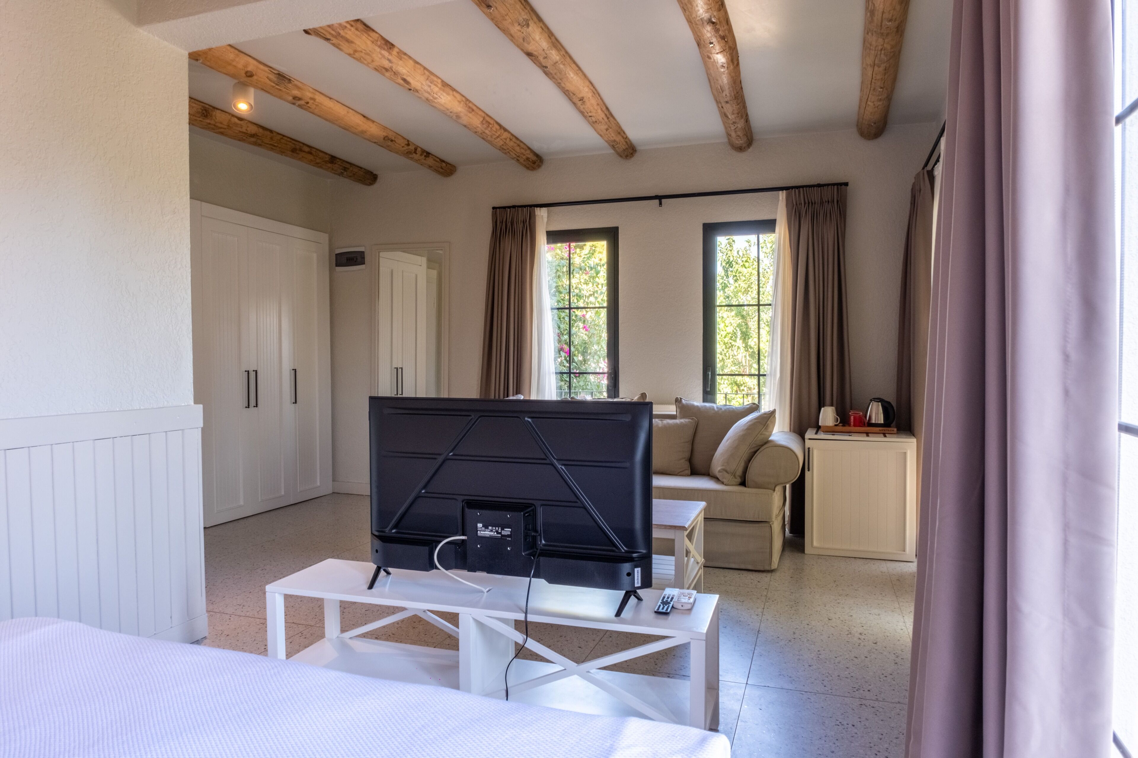 Habitació moderna amb un llit, una televisió sobre un suport blanc, un sofà i una taula petita amb una paperera i tasses.