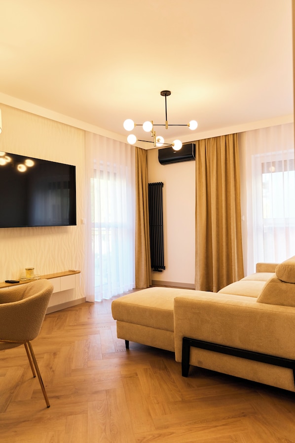 A modern living room with a sofa, a TV mounted on the wall, a wooden floor, and a ceiling light fixture. - BP Gołd Premium (Toruń)