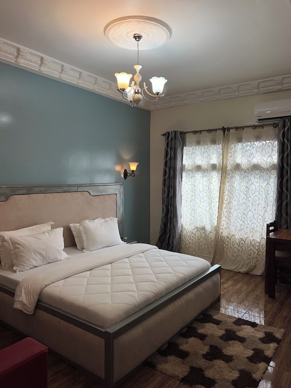 A bedroom with a bed, a nightstand, a wall-mounted lamp, a ceiling fan, a window with curtains, and a wooden floor.