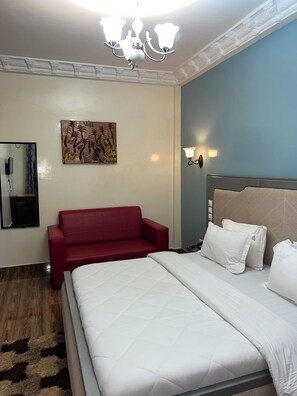 A hotel room with a bed, a red sofa, a wall-mounted artwork, a wall sconce, and a mirror.