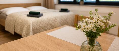 A hotel room with two beds, a wooden table with a vase of flowers, and a window with blinds.