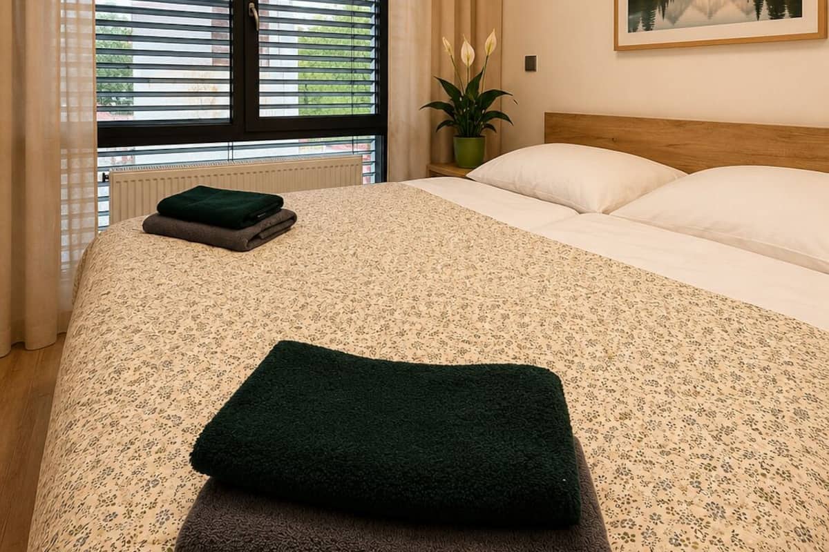 A hotel room with a bed, a window with blinds, a painting on the wall, and a radiator.