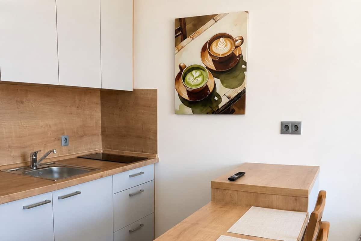 A modern kitchen with a wooden countertop, a sink, and cabinets. There is a small table with two chairs and a painting of coffee cups on the wall.