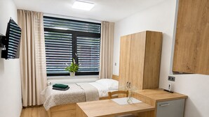 A compact hotel room with a bed, a small desk, a chair, a wooden wardrobe, a window with blinds, and a potted plant. - Microbyty & Apartments  (Ústecký kraj)