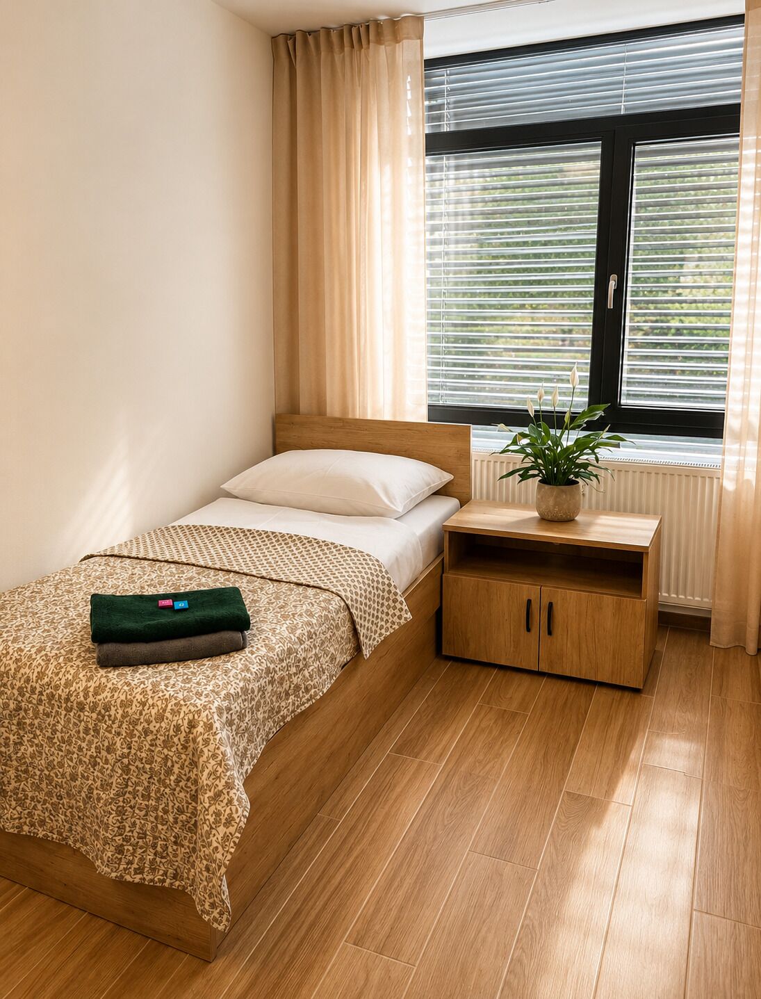 Ein Einzelbett mit gemustertem Überwurf, ein Nachttisch mit Pflanze und ein Fenster mit Jalousien.