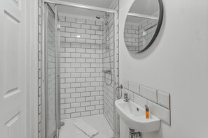 Un baño moderno con cabina de ducha de vidrio, paredes con azulejos blancos, un espejo redondo y un lavabo blanco con una toalla.