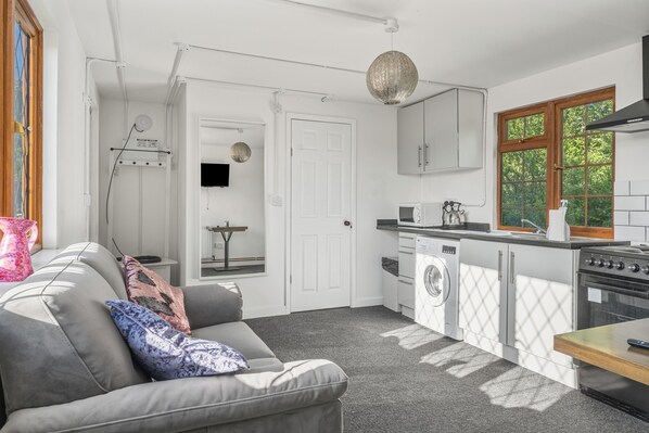 A compact kitchen with a washing machine, a dining area with a table, and a grey sofa with patterned cushions.