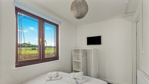Quarto com uma janela grande, cama com toalhas, guarda-roupa branco e televisão.