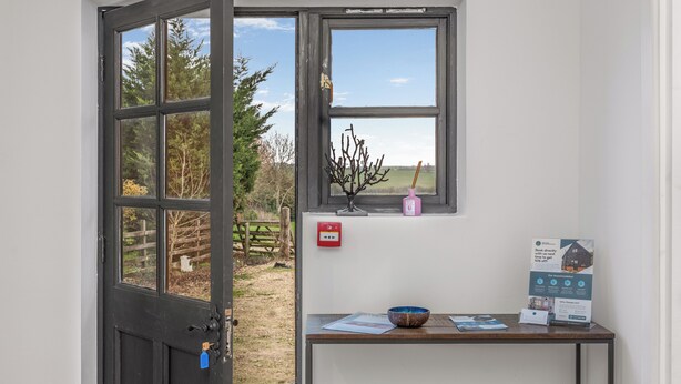 Une pièce avec une grande fenêtre, une table en bois sur laquelle se trouvent un bol et des papiers, et une porte avec une clé.