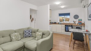 Un salon moderne avec un canapé vert, une table à manger avec une chaise, et un coin cuisine doté de meubles blancs et d’une fenêtre.