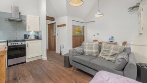 A compact kitchen with a stove, oven, and white cabinets. There is a grey sofa with plaid cushions in a living area. - The Nook - Cosy Rural Cottage with Stunning Views (Charndon)