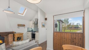 A cozy living room with a fireplace, a dining table, and a kitchen in the background. - The Nook - Cosy Rural Cottage with Stunning Views (Charndon)