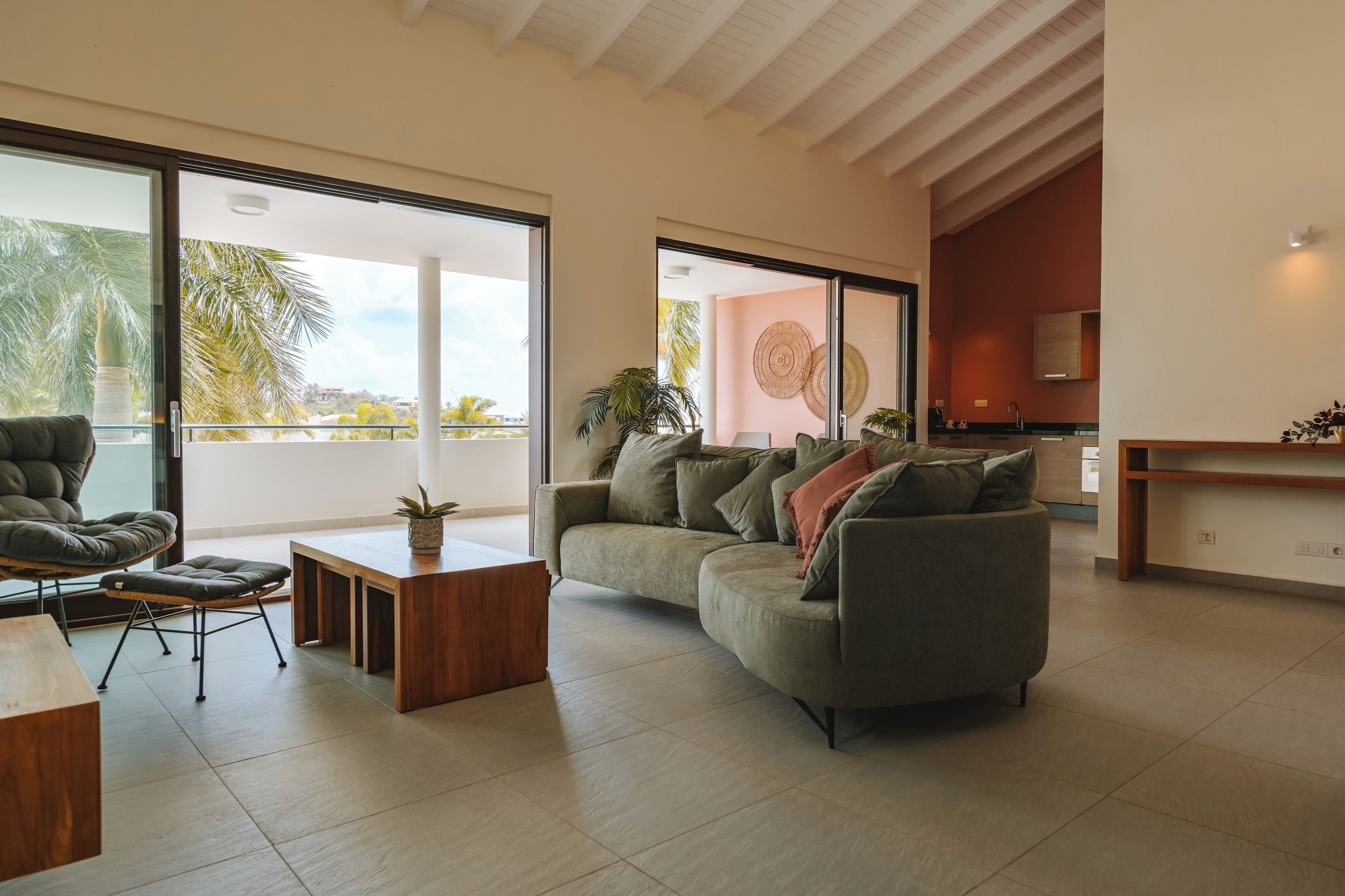 Una sala de estar moderna con un sofá, una mesa de centro y una silla. Se ve un ventanal con vistas a palmeras.