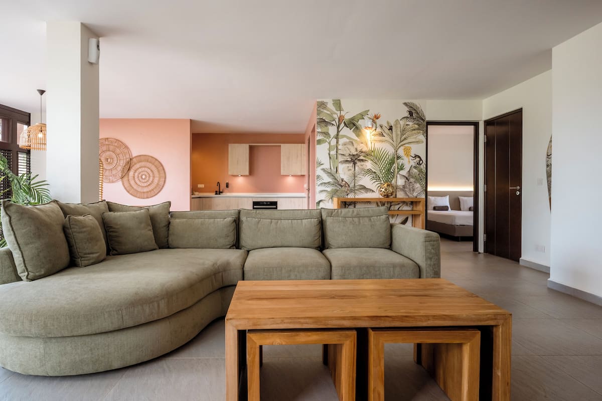 A living room with a sectional sofa, a wooden coffee table, a kitchen in the background, and a tropical-themed wall art piece.
