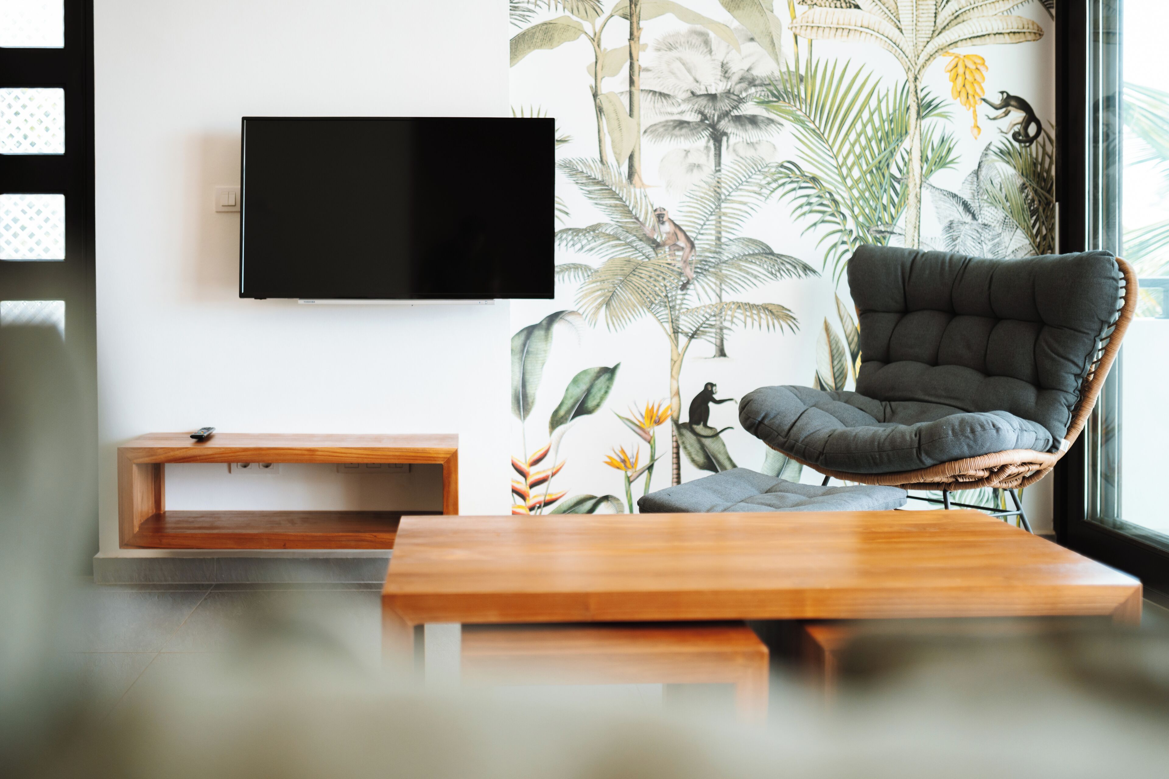 A modern living room with a flat-screen TV, a cushioned chair, a wooden coffee table, and a wall with a tropical plant mural.
