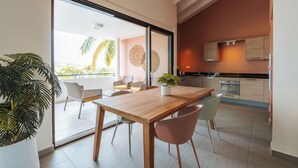 Una cocina moderna con armarios de madera y una isla grande, un comedor con mesa y sillas de madera, y un amplio ventanal corredero que da a un balcón con vistas a palmeras.
