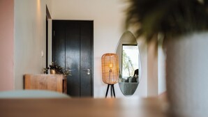 Un interior moderno con una puerta oscura, una lámpara de mimbre y un espejo redondo que refleja una planta y un sofá.
