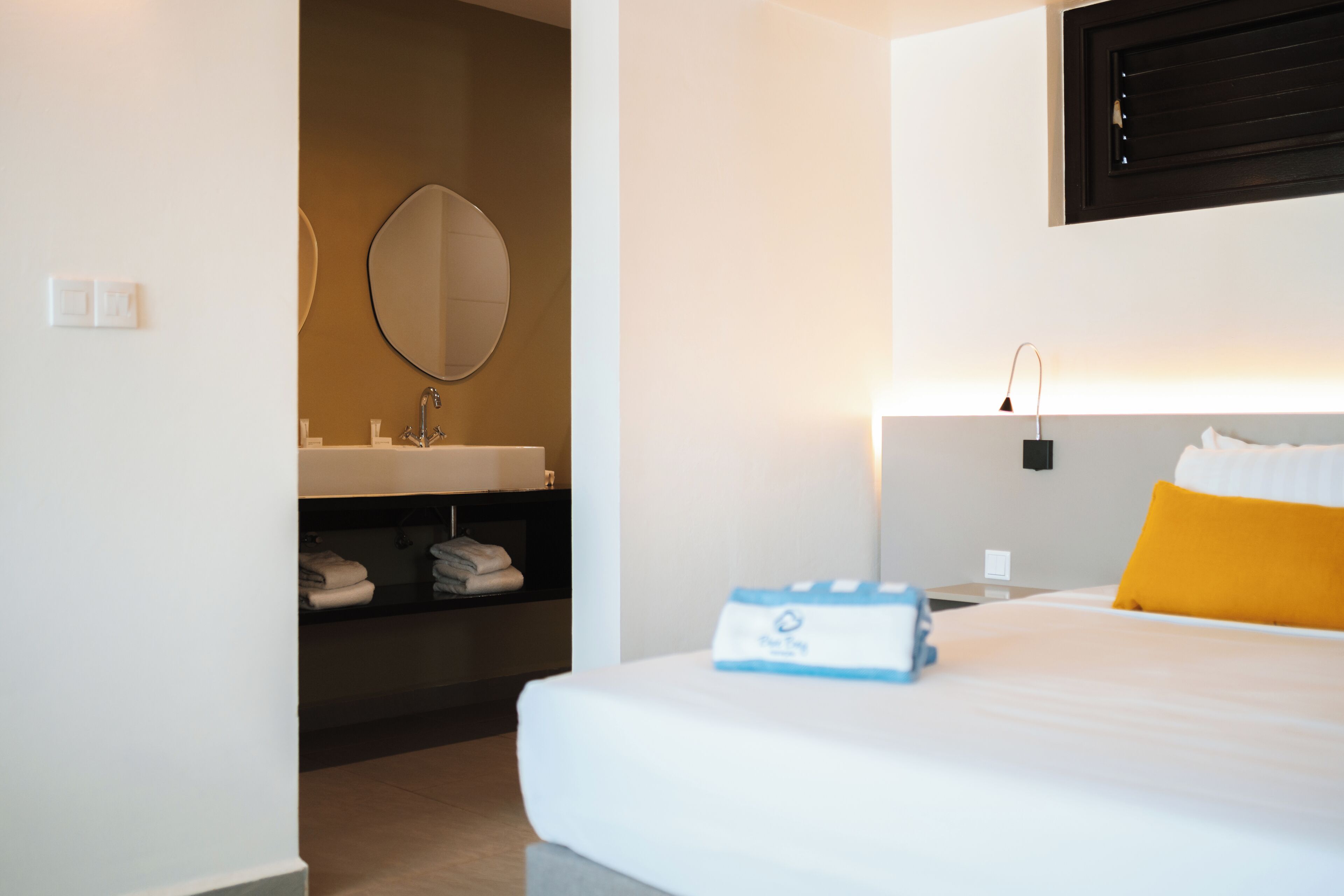 A hotel room with a bed, a bedside table, a nightstand, a wall-mounted mirror, a bathroom with a sink, and a towel rack.