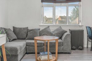A corner room with a grey-striped sofa, a round wooden table with books, a radiator, a window with blinds, and a trash bin. - Flat B: Buckingham Road (Oxfordshire)