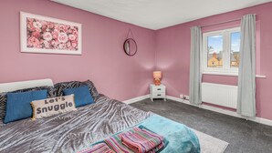 Un dormitorio con cama, almohadas, una obra de arte floral enmarco, una mesita de noche con lámpara y una ventana con cortinas.