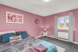 A bedroom with a bed, pillows, a framed floral artwork, a bedside table with a lamp, and a window with curtains. - Fox Haven - Charming Three-Story Cottage in Bicester (Bicester)