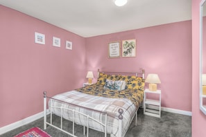 A bedroom with a bed, two bedside tables, a headboard, a pink wall, and framed pictures.