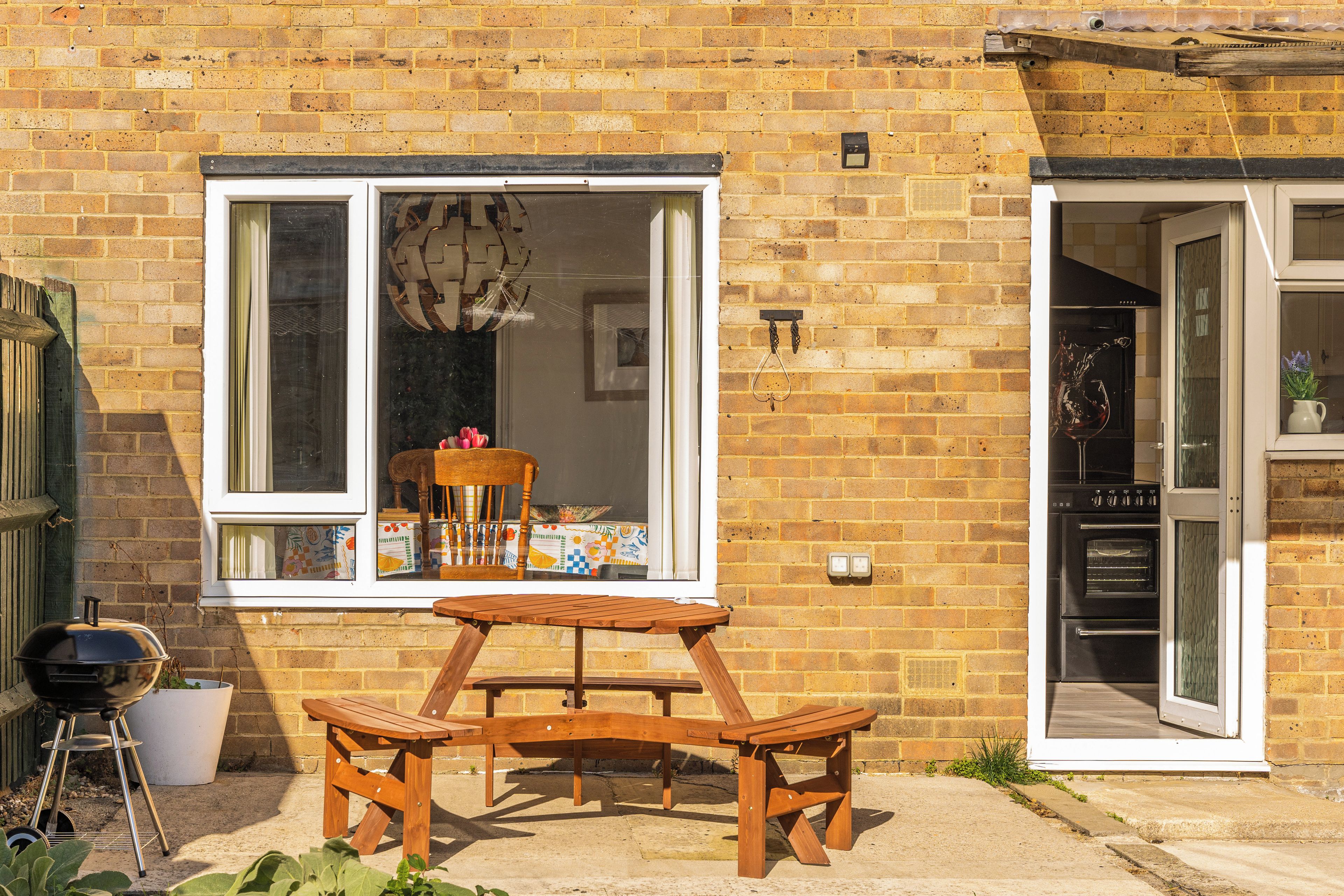 A brick house with a patio table and chairs, a wooden chair inside, and a grill outside.
