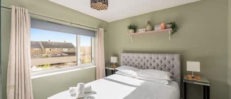 A hotel room with a bed, a bedside table, a nightstand, a window with curtains, a hanging light fixture, and a view of buildings outside.