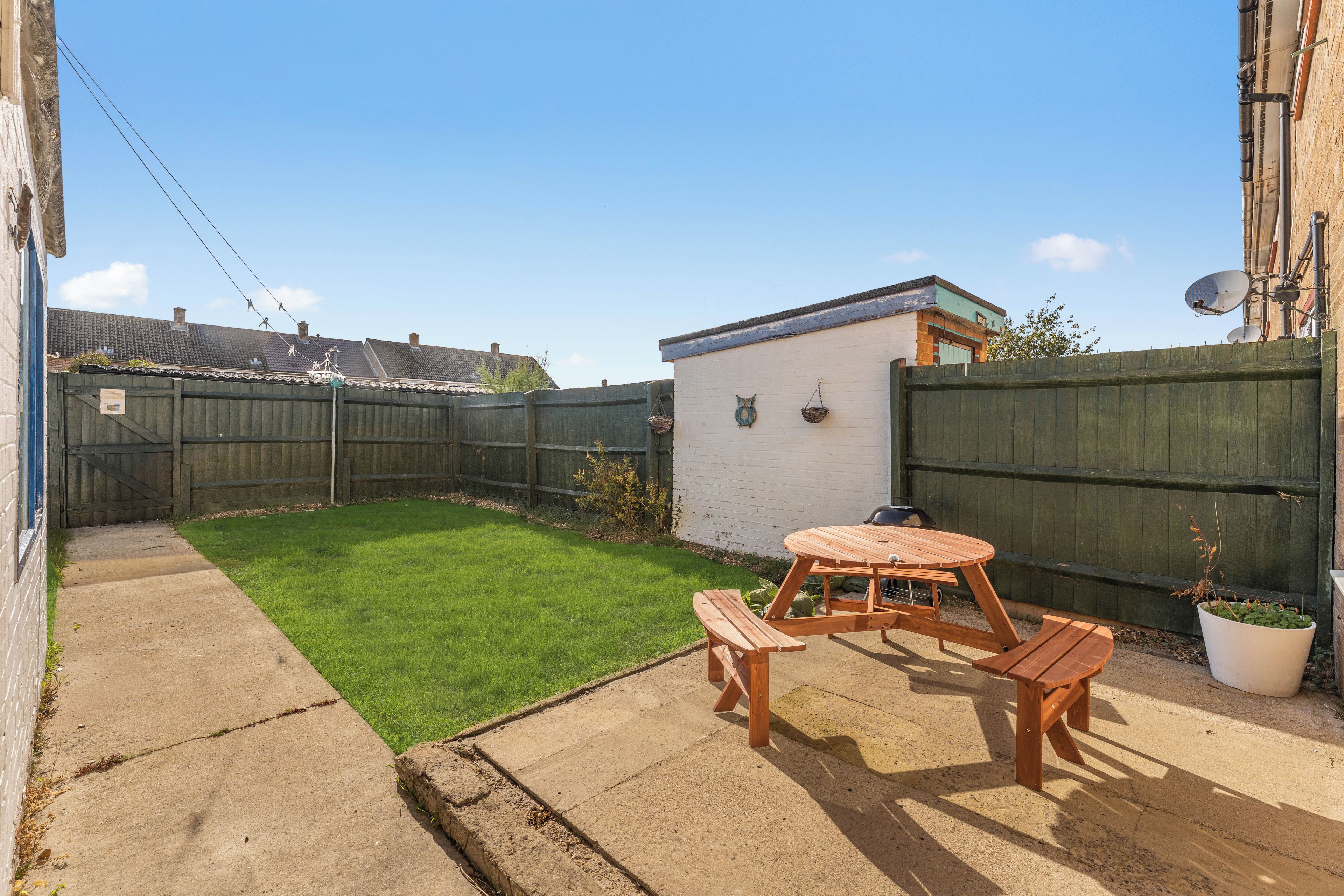 A backyard with a wooden table and benches, a small white shed, and a fenced area.
