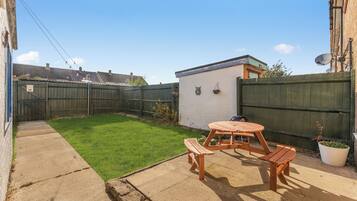 A backyard with a wooden table and benches, a small white shed, and a fenced area.