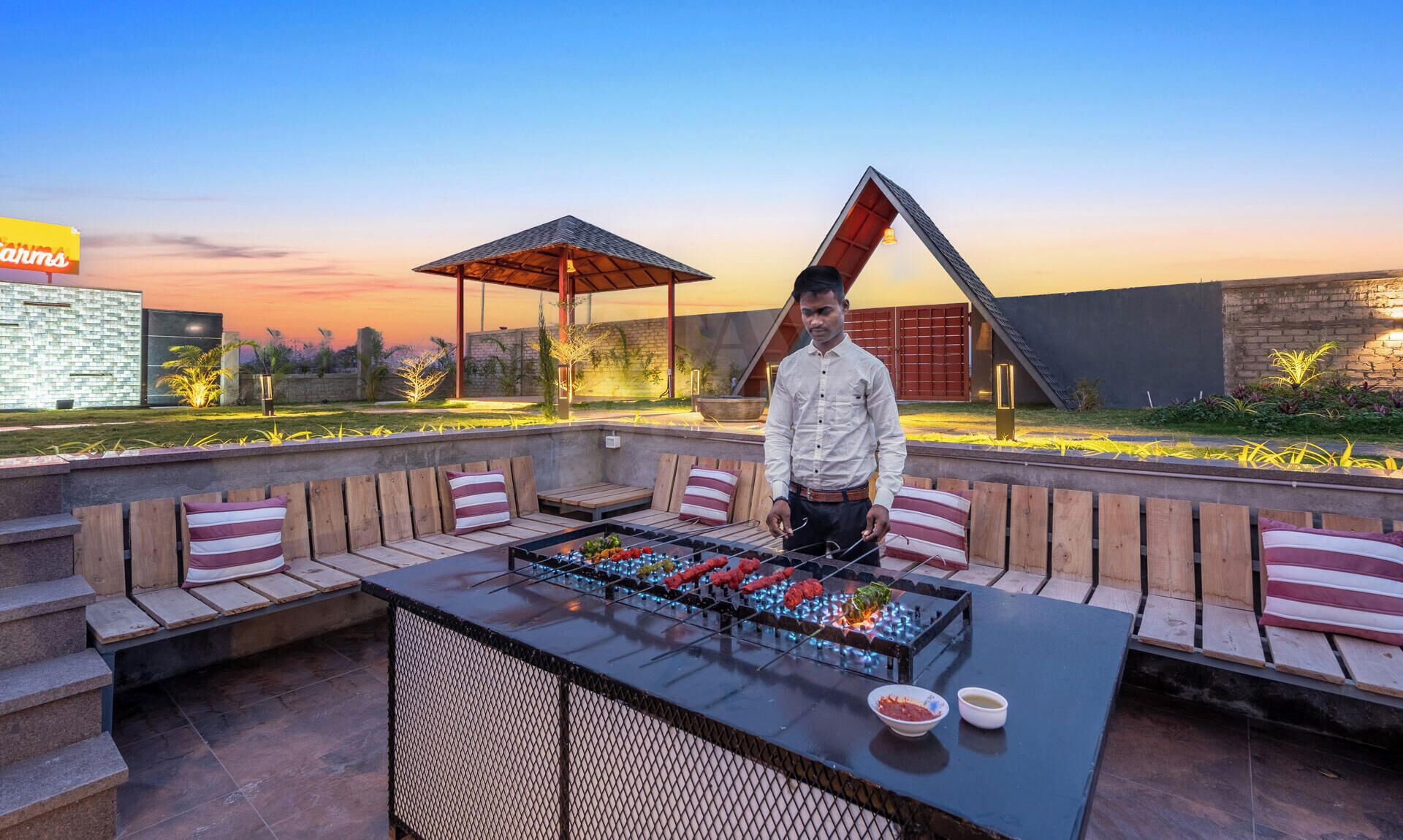 A man standing by a barbecue grill with seating and a covered area, set against a sunset backdrop.
