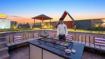 A man standing by a barbecue grill with seating and a covered area, set against a sunset backdrop.