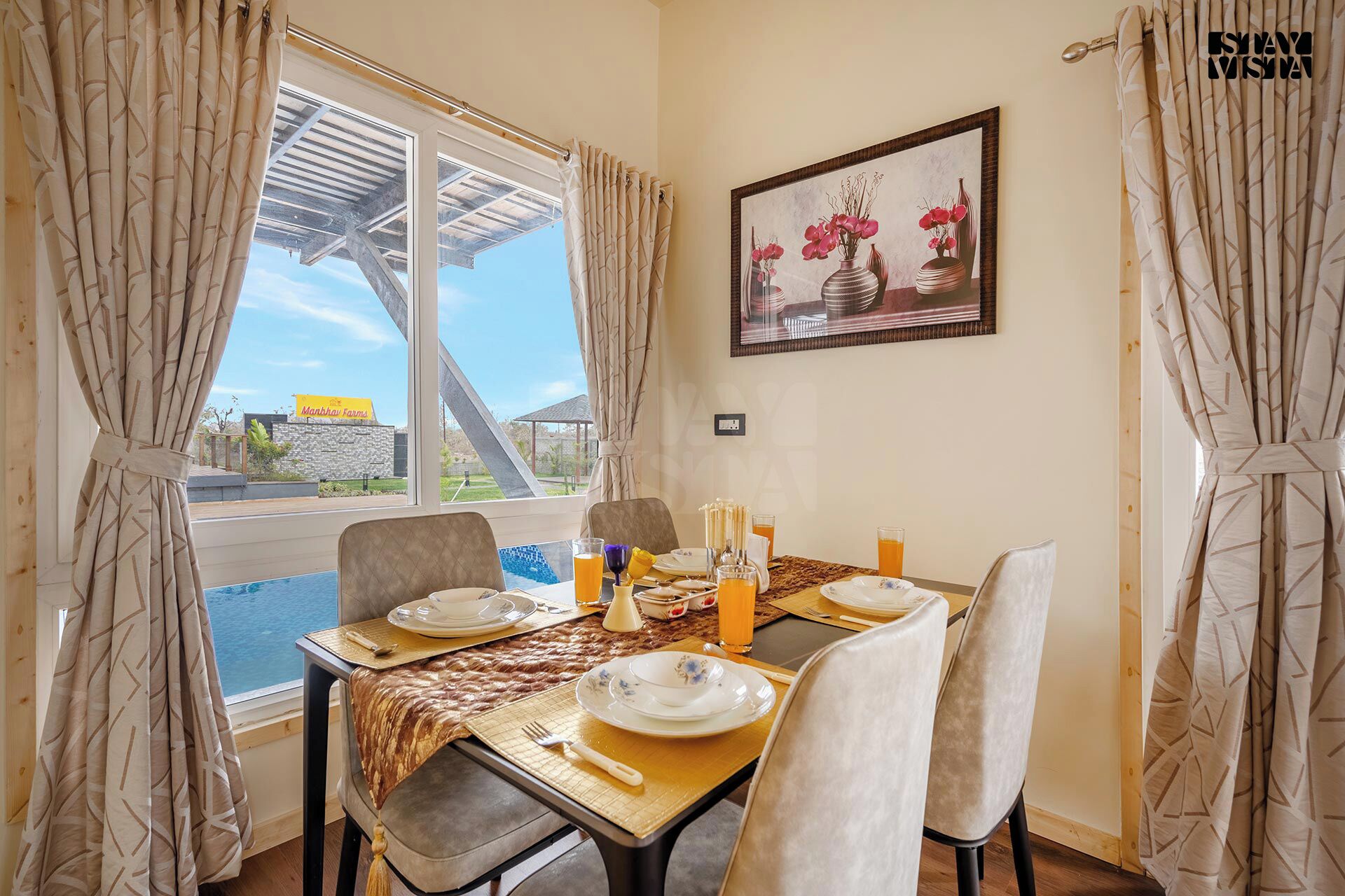 Un rincón comedor con una mesa dispuesta para dos, un cuadro en la pared, y vista a una piscina y un edificio.