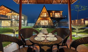 Una casa moderna con techo triangular, balcón y comedor con muebles de mimbre y mesa lista para dos comensales.