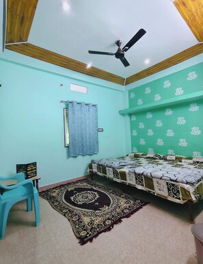 A bedroom with a bed, a ceiling fan, a patterned rug, a blue chair, and a wall-mounted curtain. - Kavya Homestay  (Ujjain)