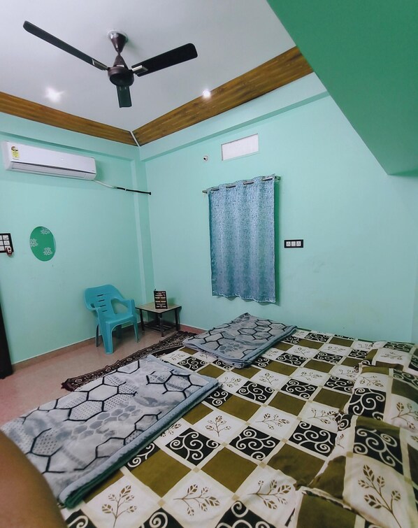A room with a bed, a ceiling fan, an air conditioner, a blue plastic chair, and a small table. - Kavya Homestay  (Ujjain)