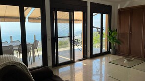 A spacious living room with large sliding glass doors leading to an outdoor patio with a table and chairs, a view of the sea, and a wooden cabinet.