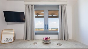 A room with a TV mounted on the wall, a window with curtains, a table with a mirror, a bowl of flowers, and a wicker chair. - Paros Golden Beach flats Casa Costa Azor - 33m from beach, sleeps 6 (3BDR 2BA) (Paros)