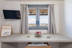 Ein Zimmer mit einem an der Wand befestigten Fernseher, einem Fenster mit Vorhängen, einem Tisch mit Spiegel, einer Blumenvase und einem Korbstuhl.
