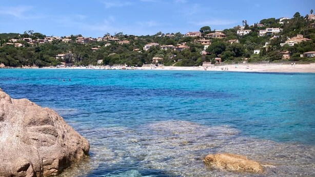 Un paysage côtier avec une eau claire, des rochers et une plage éloignée, où l’on aperçoit des bâtiments.