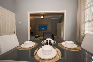 Ein Esstisch ist gedeckt mit weißen Tellern, Schüsseln und Besteck, im Hintergrund ist ein Fernseher zu sehen.
