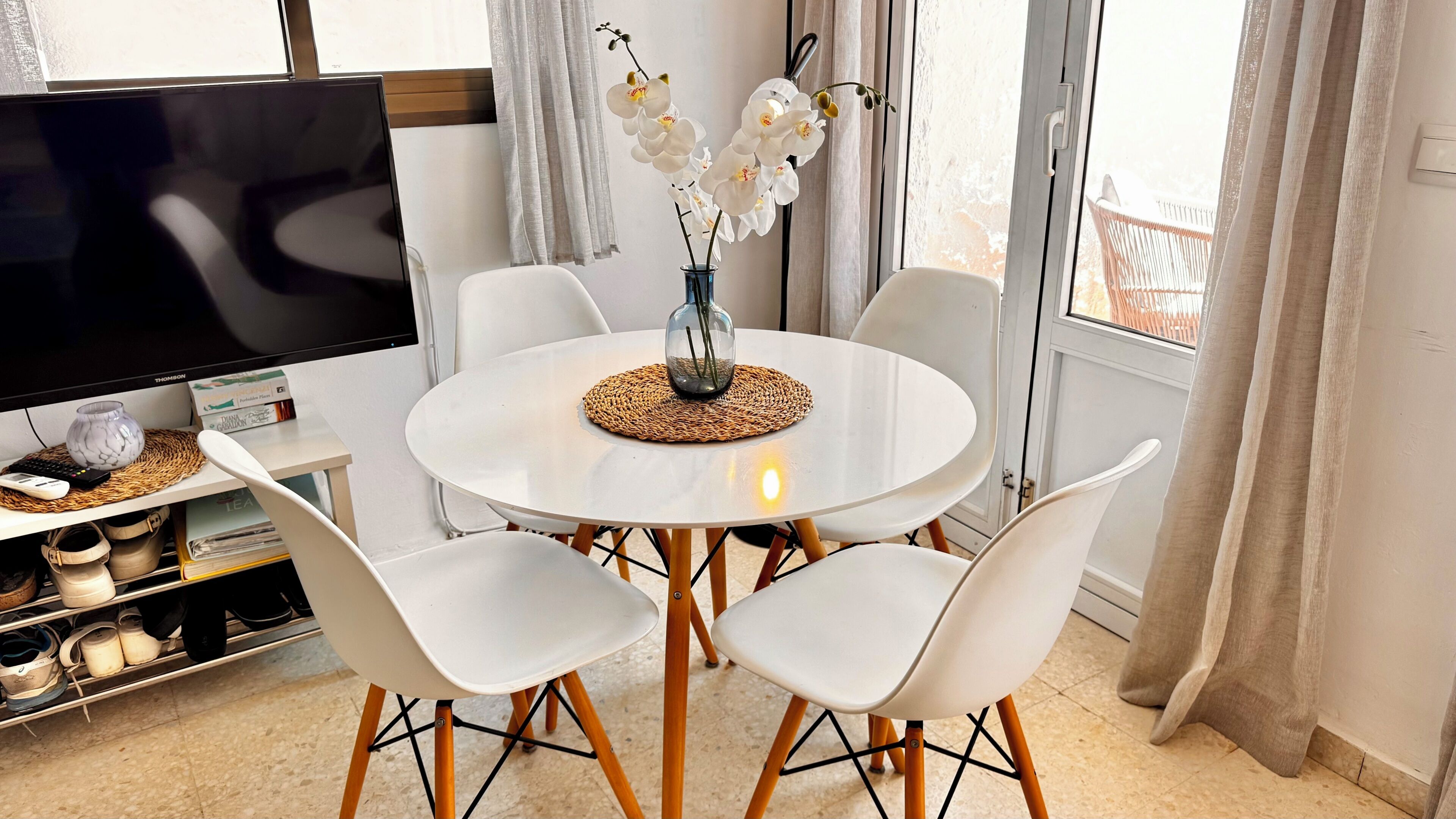Uma sala de jantar com uma mesa redonda, cadeiras brancas, um vaso com flores brancas, uma televisão e uma estante com sapatos.