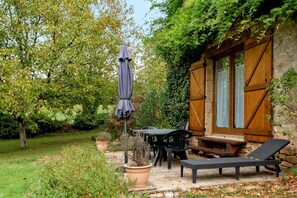 A rustic outdoor seating area with a table, chairs, and a lounge chair, surrounded by greenery and a stone wall.