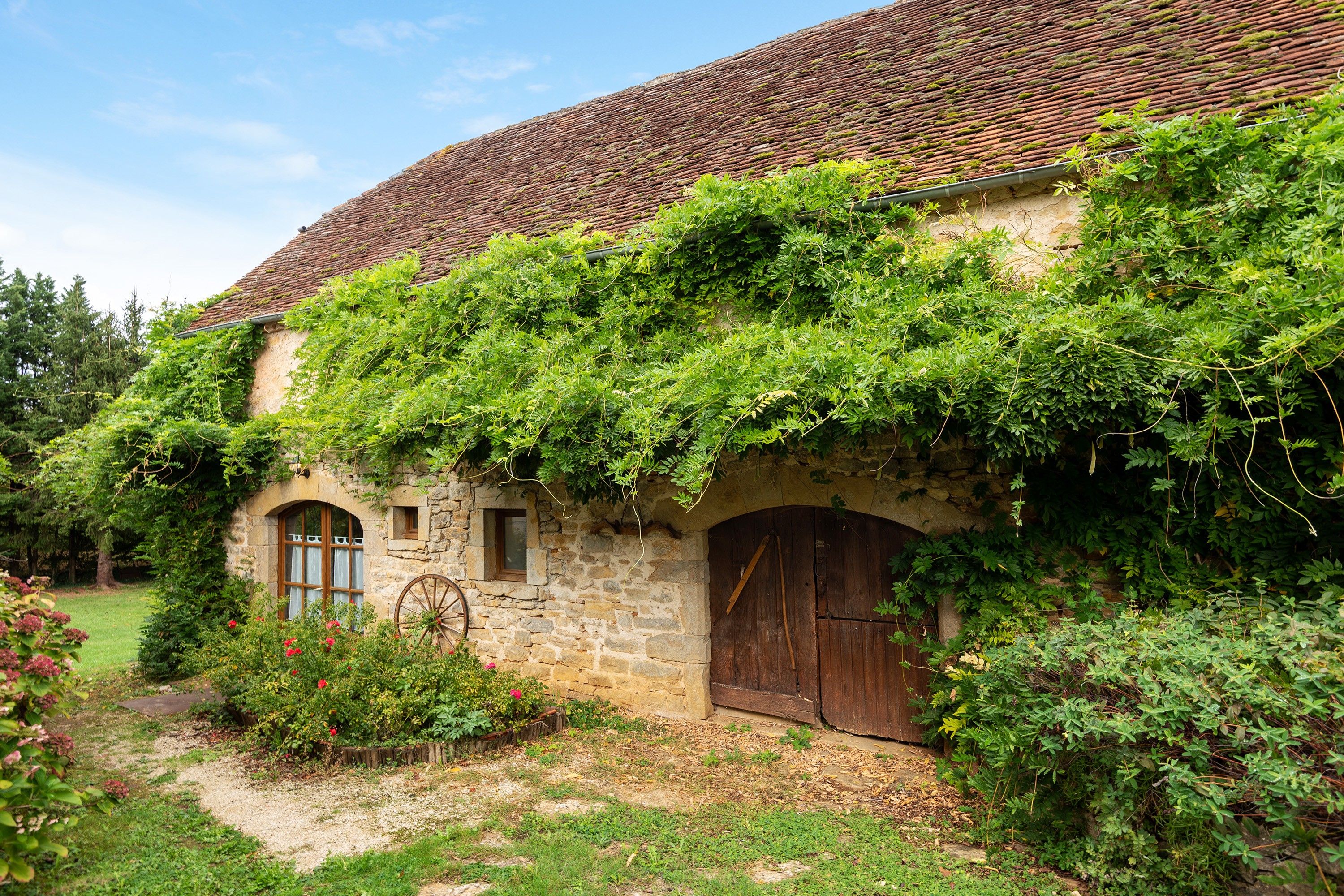 A rustic stone building with a tiled roof, a wooden door, and a wheelbarrow in the foreground.
