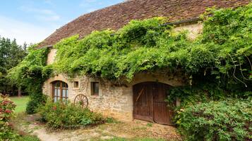 A rustic stone building with a tiled roof, a wooden door, and a wheelbarrow in the foreground.