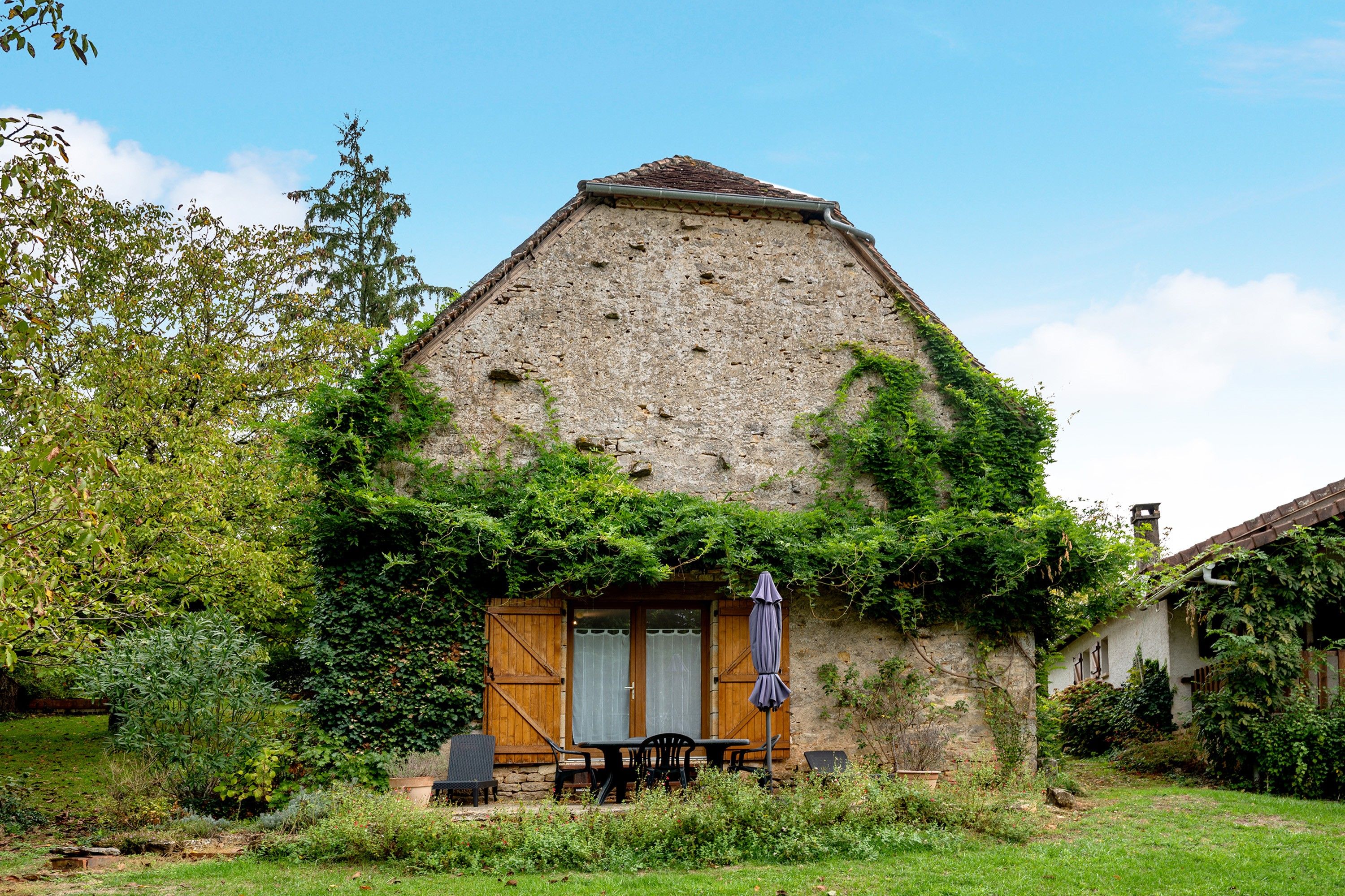 Un bâtiment rustique en pierre, doté d’un toit de chaume, d’une porte en bois et d’un petit espace extérieur aménagé avec une table et des chaises.