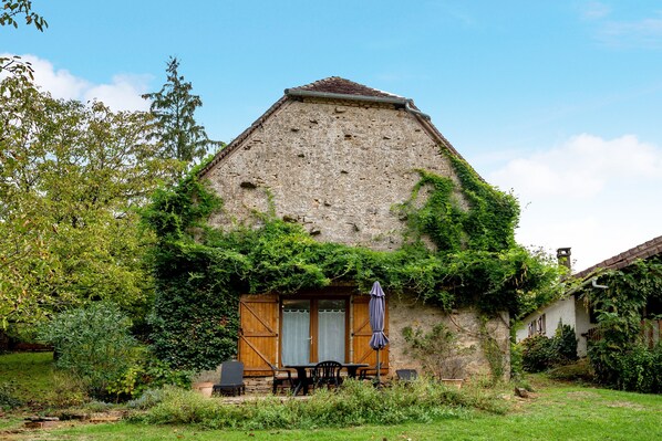 A rustic stone building with a thatched roof, a wooden door, and a small outdoor seating area with a table and chairs.
