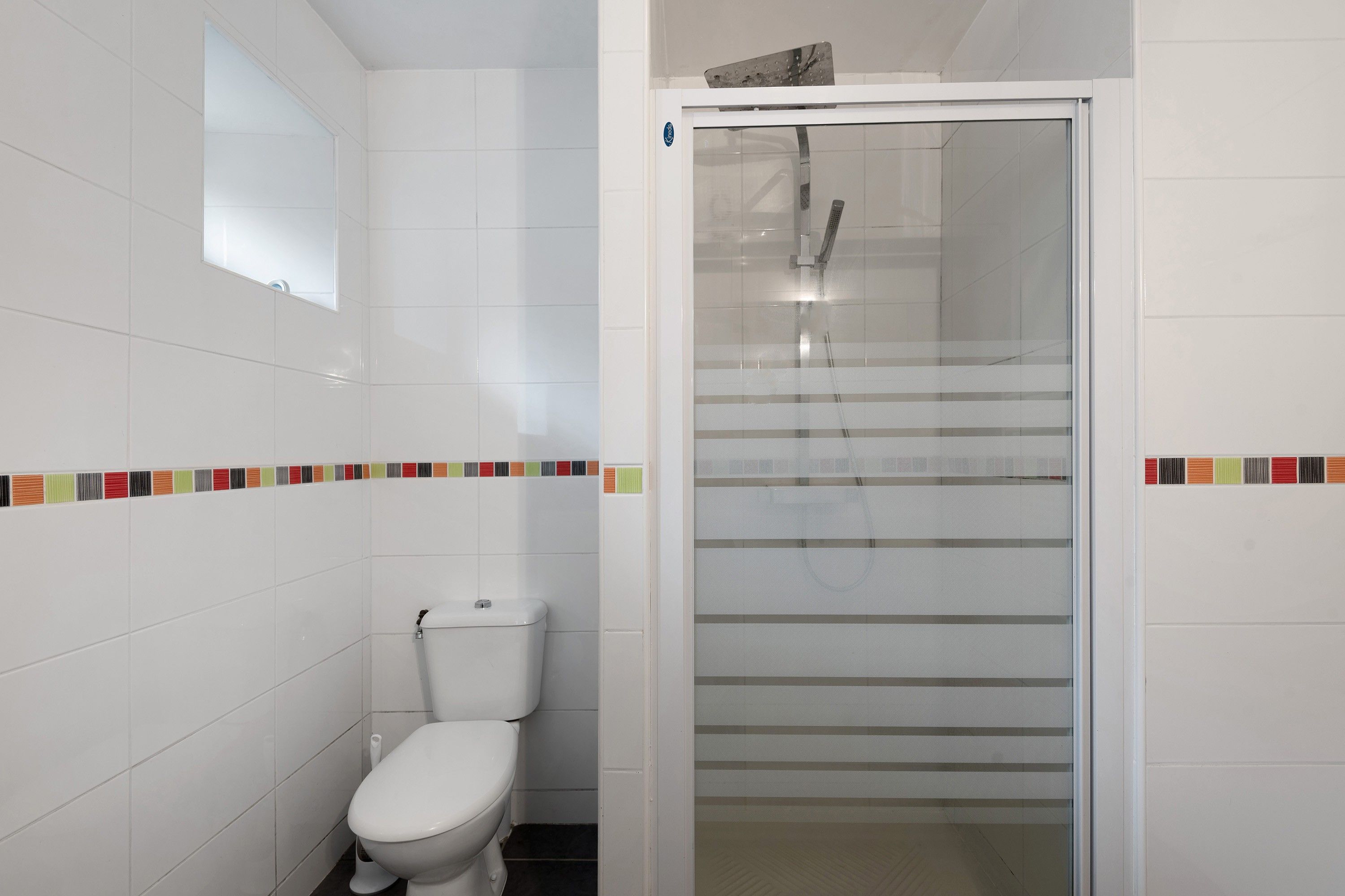 Une salle de bain comprenant des toilettes, une douche avec une porte en verre et un mur carrelé agrémenté d’une bordure colorée.