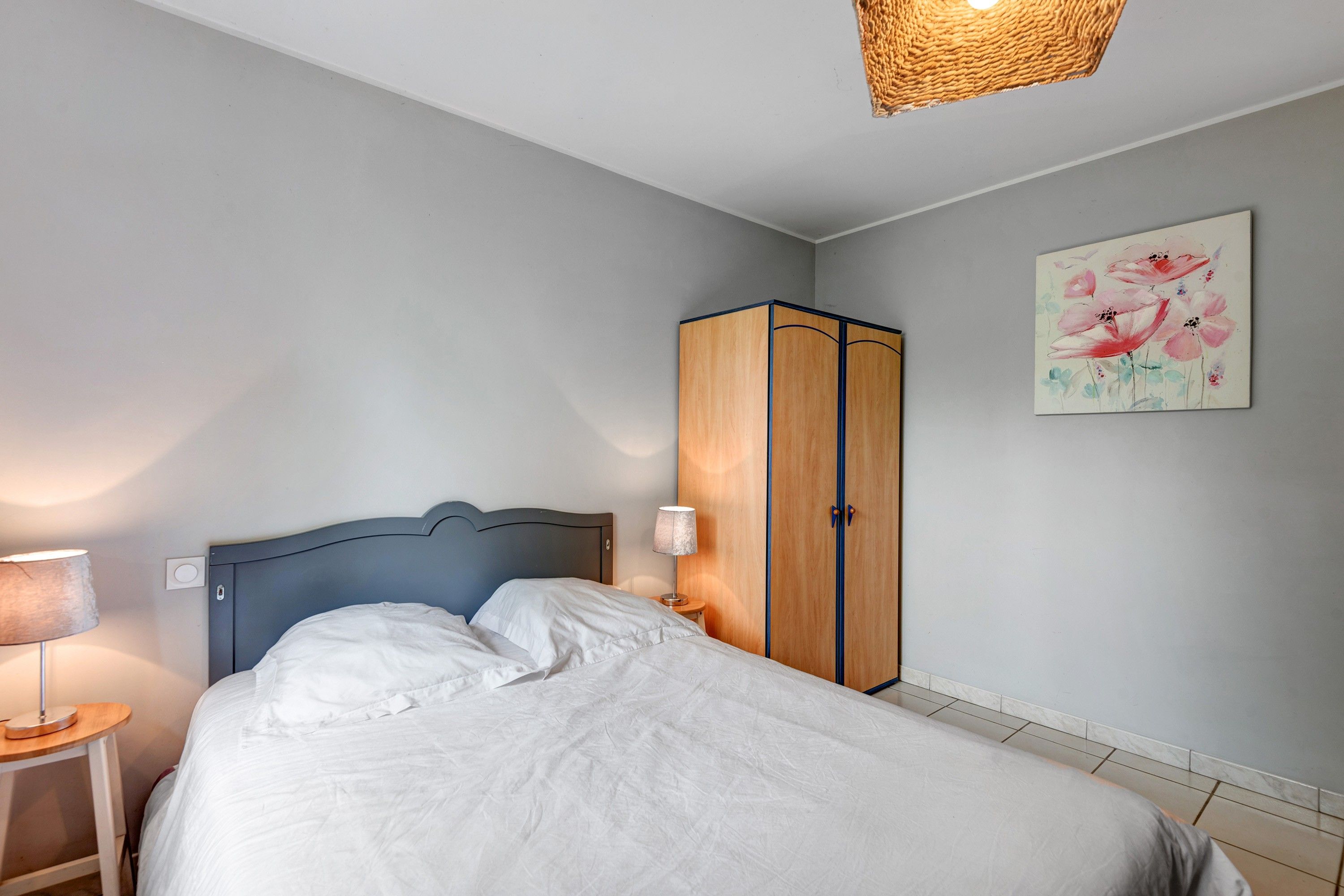 A bedroom with a bed, a wardrobe, a nightstand, a lamp, and a wall painting.
